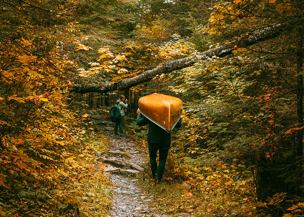 A couple hiking the trails, carrying a canoe through the fall forest.
